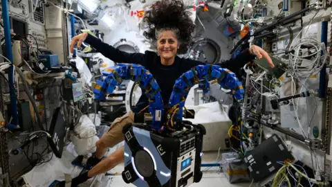 NASA A smiling Suni Williams floating in the international space station, in a black long-sleeve top and beige shorts, hair wild and having a life of its own as it floats in zero gravity. Her arms are outstretched mimicking a box-like apparatus floating in front of her which has two thick arm-like blue appendages emerging from its top and spreading out. In the background the interior walls of the ISS are a messy riot of different coloured cables, keypads and monitors.