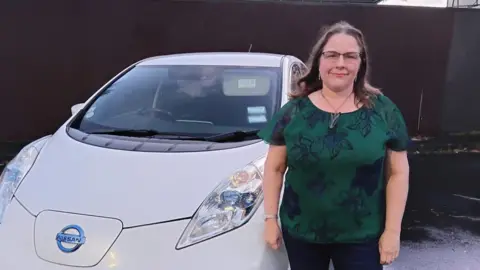 Lucy Crawford Wearing a dark green top, Lucy Crawford stands next to her white Nissan Leaf