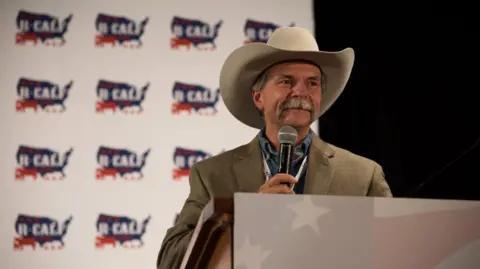 Bill Bullard A man wearing a cowboy hat speaks into a microphone.