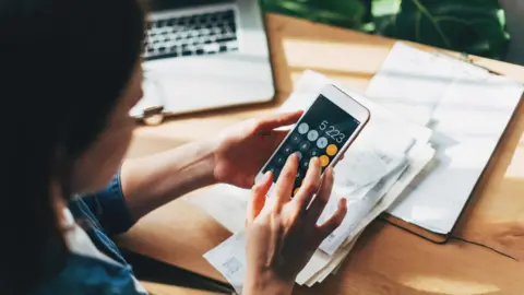 Getty Images Woman sits at a desk with paperwork and a laptop in front of her. A smartphone in her hand has a calculator on the screen.