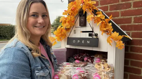 BBC Annabelle Cox carrying a tray of cookies is standing in front of her honesty box which looks like a small, white shed. There is an open sign, a slot for cash, a digital doorbell and a garland of autumn leaves in view on the box.