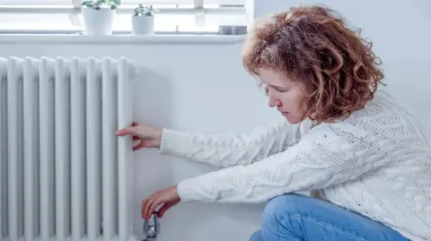 Getty Images A woman turning the dial on a radiator with one hand, while feeling the temperature with the other. She has shoulder length ginger hair and is wearing a white knitted jumper and blue jeans.
